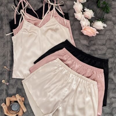 Three sets of satin camisole tops with tie-up shoulder straps and matching shorts in ivory, blush pink, and black, laid on a soft textured background with flowers.